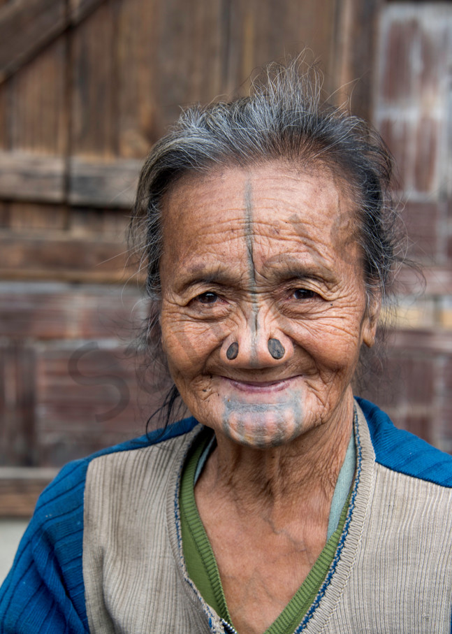 A smiling older Apatani woman with nose plugs and facial tattoos
