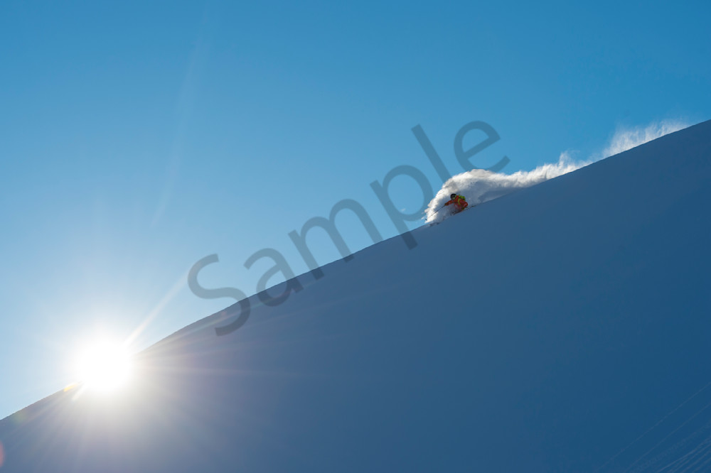 Salomon skier Brody Leven skiing steep turns down a snow covered slope with blue skies above and the sun shining over the ridgeline in the mountains of Whistler Blackcomb British Columbia Canada