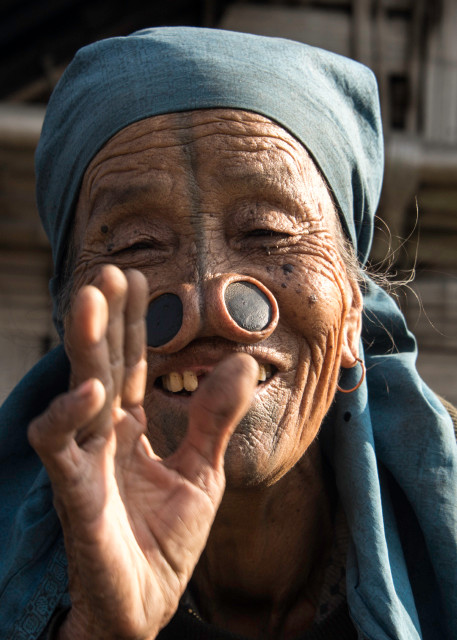 Apatani woman with large nose plugs, smiling and waving.