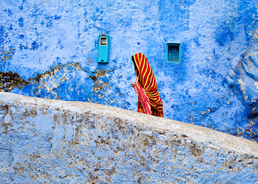 Travel photograph of woman in bright red and yellow chador walking in front of blue wall in Chefchaouan Morocco the blue city