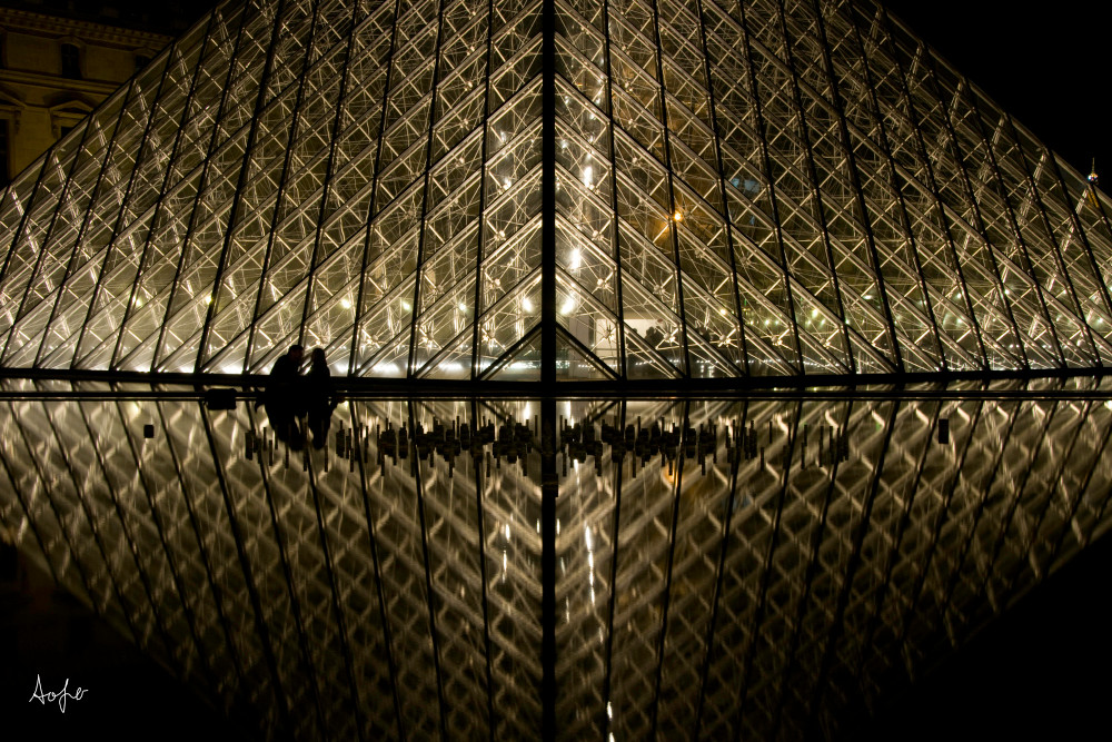 Photograph of silhouetted couple by louvre art museum at night with reflection in water