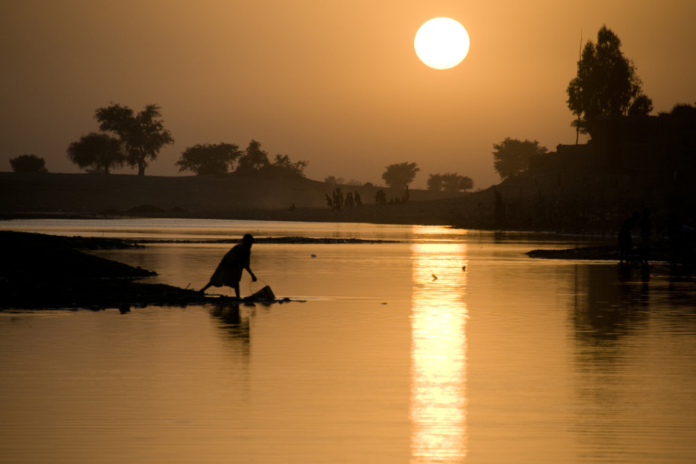 Fine art photograph of a silhouette of woman with bucket by Bani river at sunset
