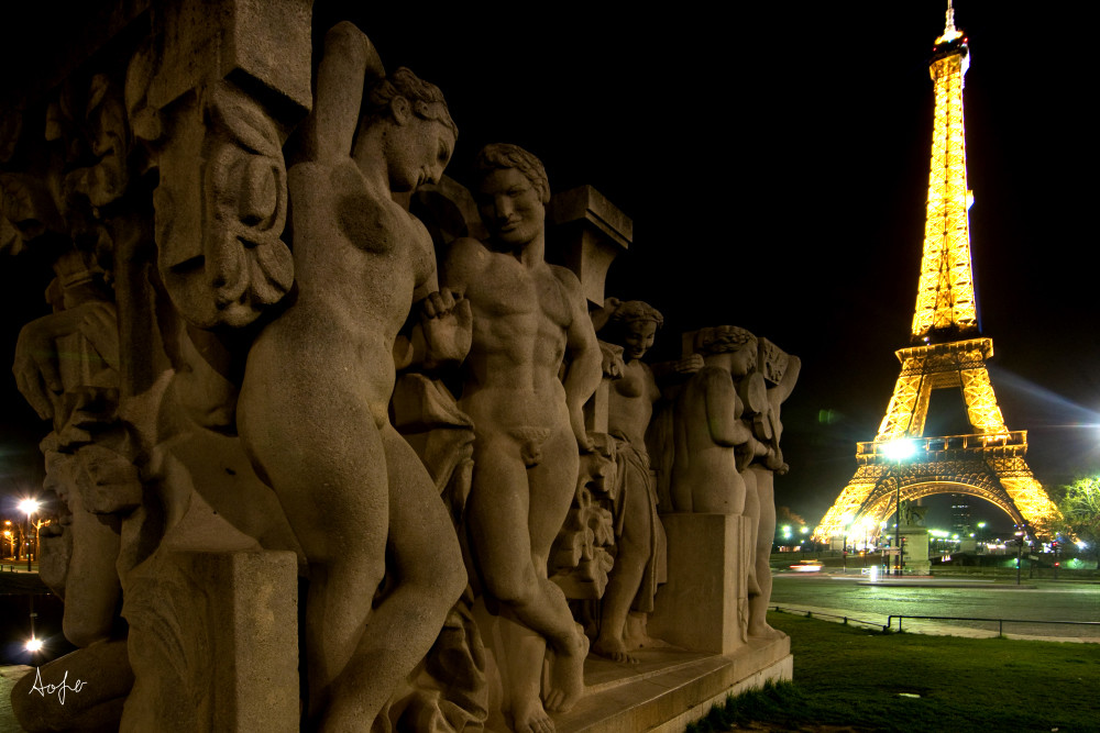 Statue of men and women at night with Eiffel tower lit up behind, in fine artphotograph