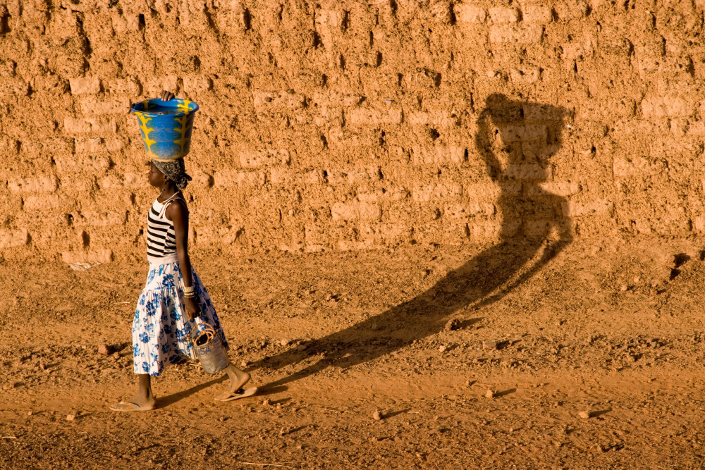 Girl with jug on head casts long shadow on mud wall, in fine art photograph 