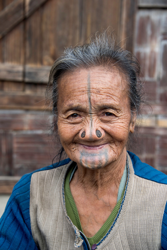 A smiling older Apatani woman with nose plugs and facial tattoos