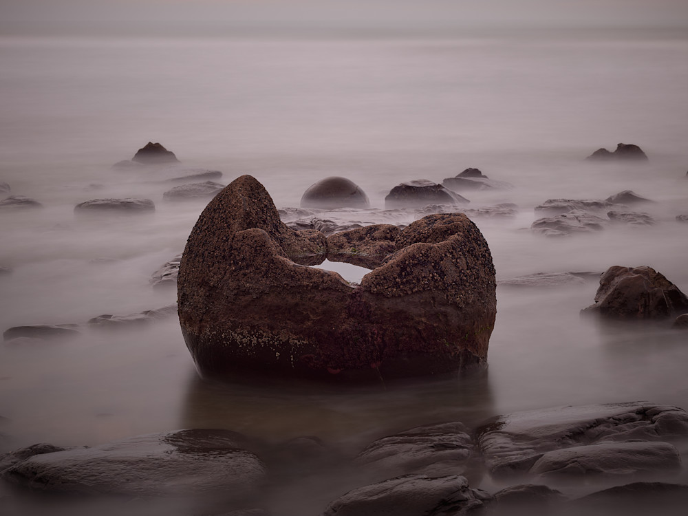 A long exposure landscape photograph of a partially submerged boulder on Moeraki beach with a pool of water in the center.