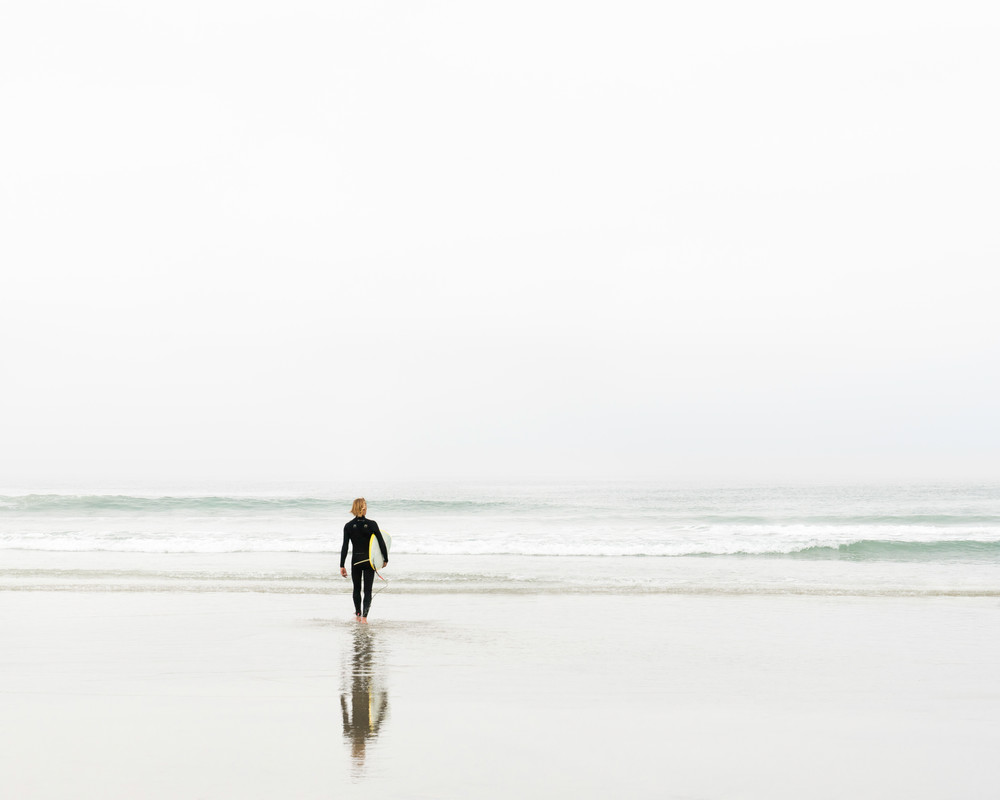 A fine art photograph of a surfer holding a surfboard looking out at the waves.
