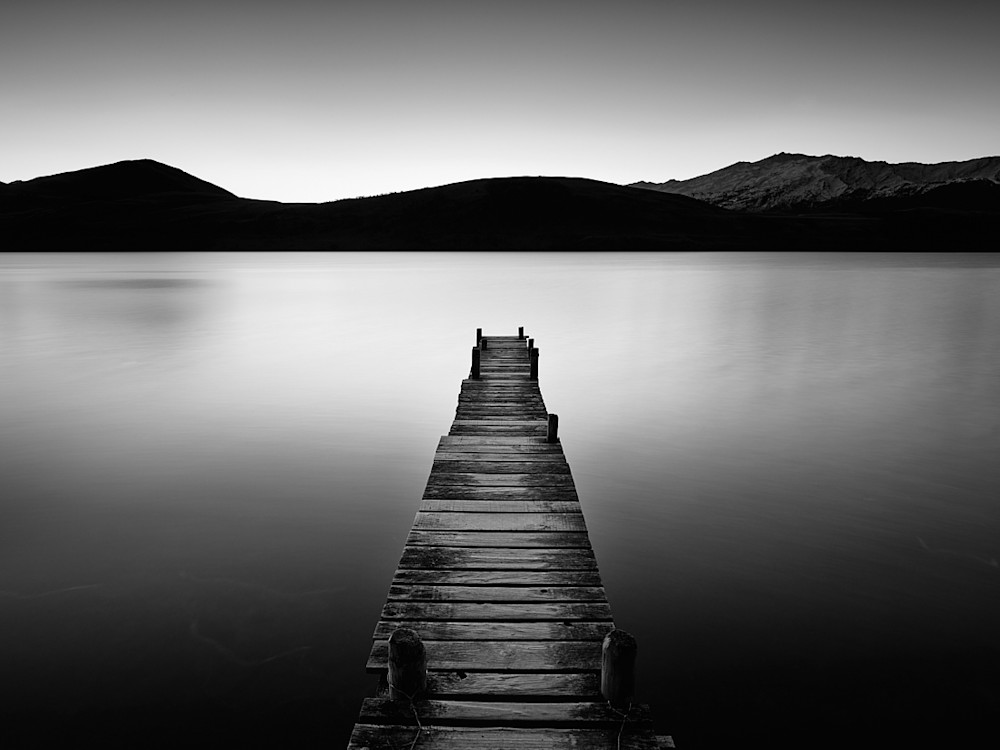 A black and white winter photograph of a lake jetty with mountains in the background.