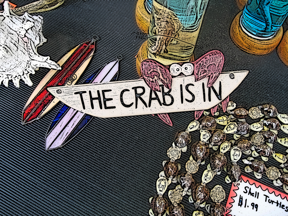 "A whimsical crab-shaped sign reads ""The Crab Is In"" among shell turtles and stained glass surfboards for sale on a table - digitally altered"