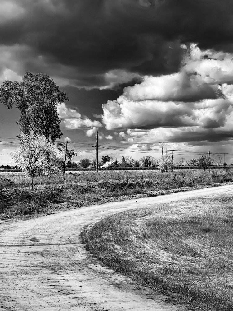 A curved dirt path marks the edge of a farm field beneath storm clouds.