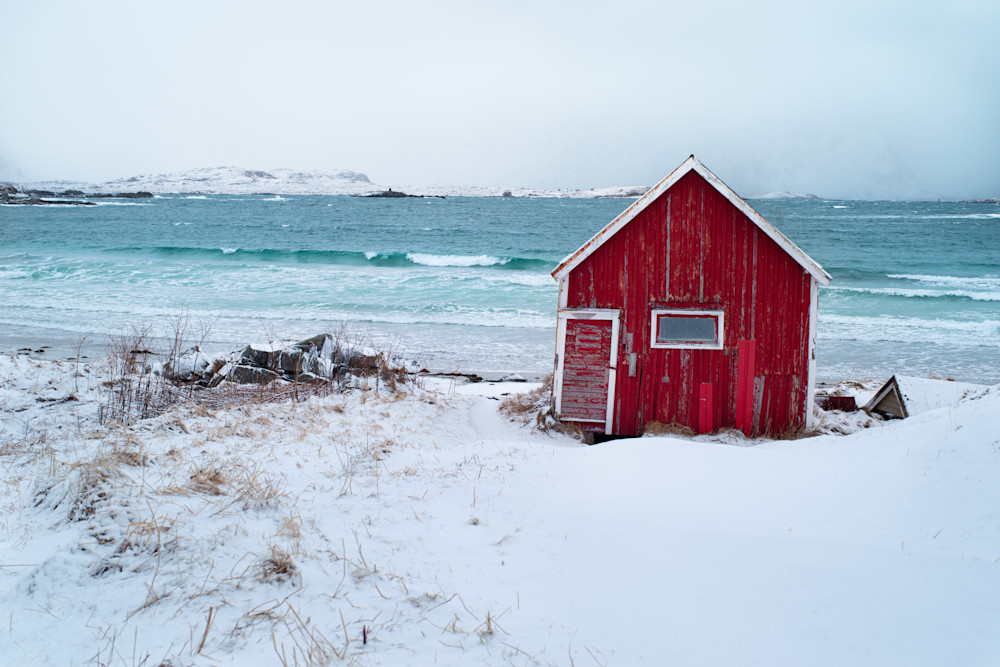 A red hut clings to the shore of a snowy beach in Ramberg on the Lofoten Archipelago, Norway