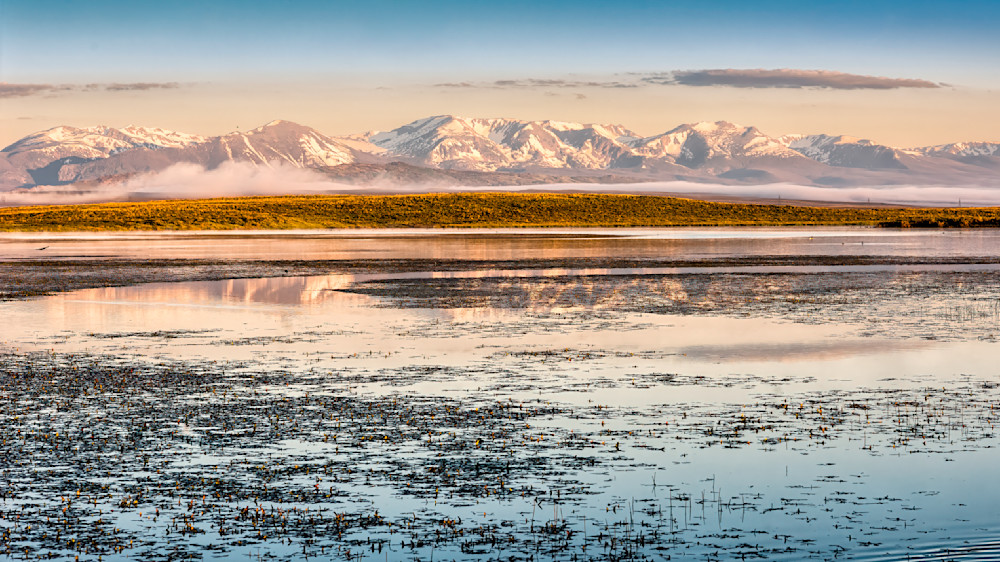 Mountainous landscape with snow-capped peaks reflecting in calm waters at dawn in a rural setting