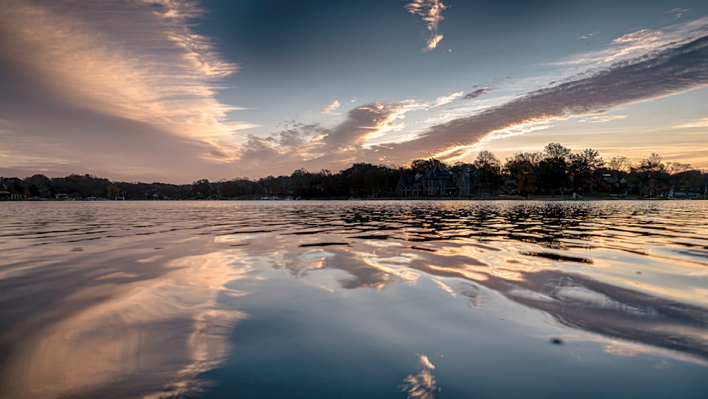 Vibrant sunrise reflecting on calm water with scattered clouds and silhouette of trees along the shore