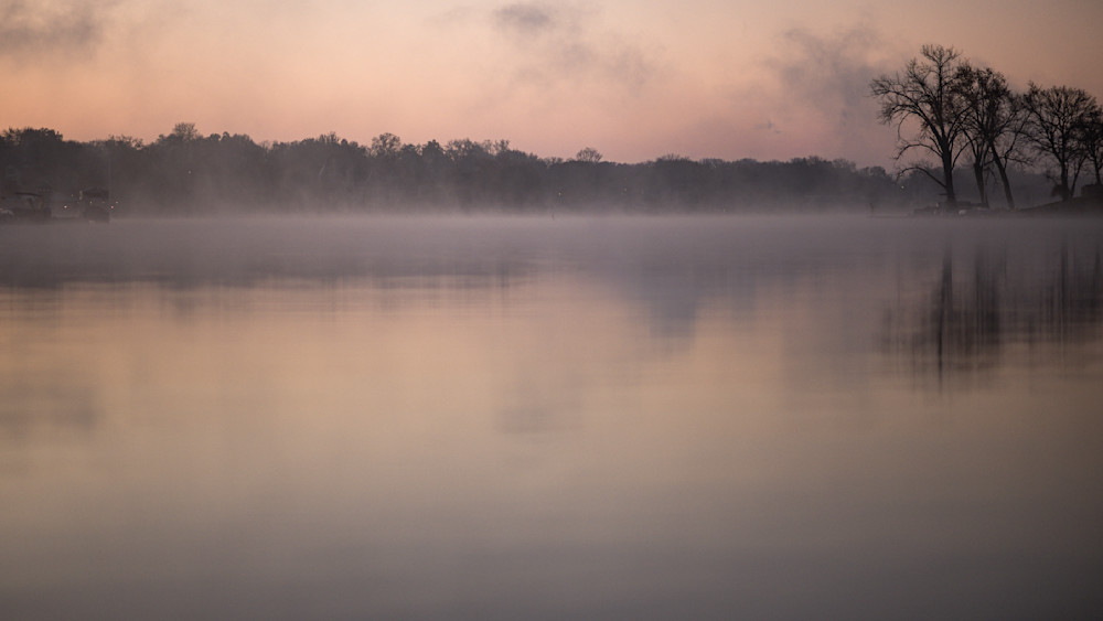 Misty sunrise over a calm lake with silhouettes of trees and soft fog covering the water surface