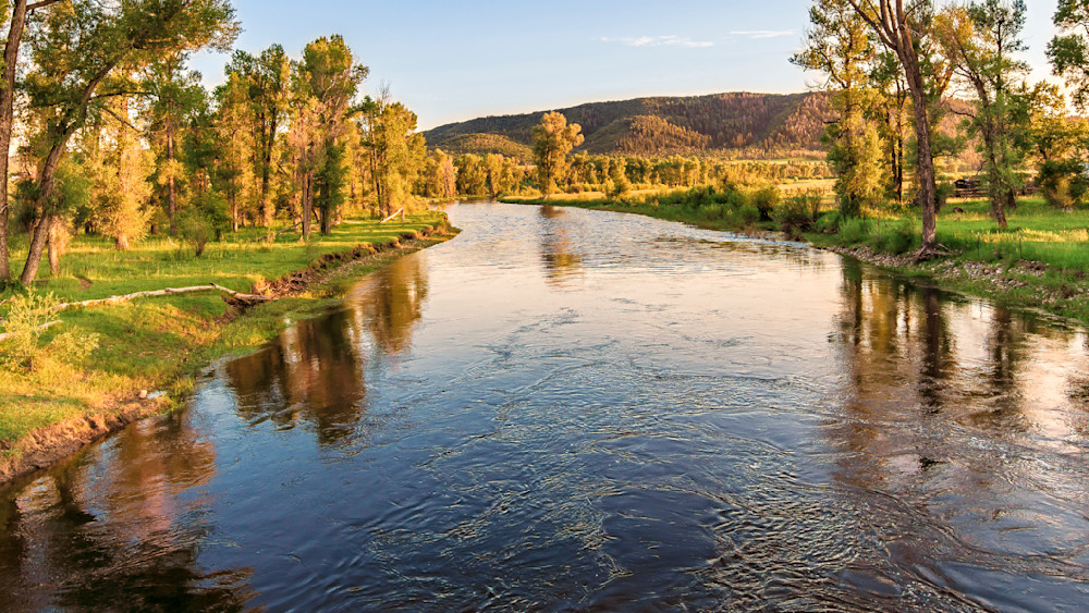 Golden hour on a river surrounded by trees and mountains near a grassy shore with clear water and blue sky