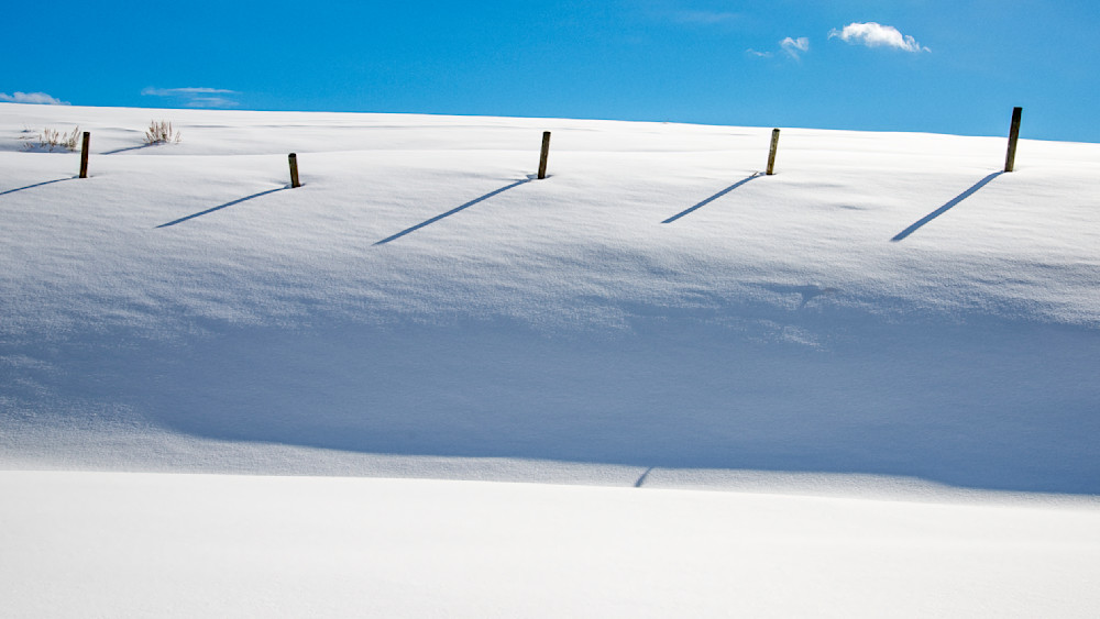 Snow line with snow-covered ground and shadows of fence posts under blue sky