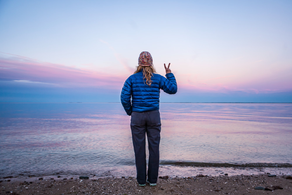 A woman standing at waters edge making a peace sign with her hand / fingers.