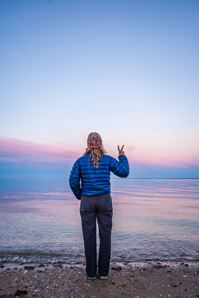 A woman standing at waters edge making a peace sign with her hand / fingers.