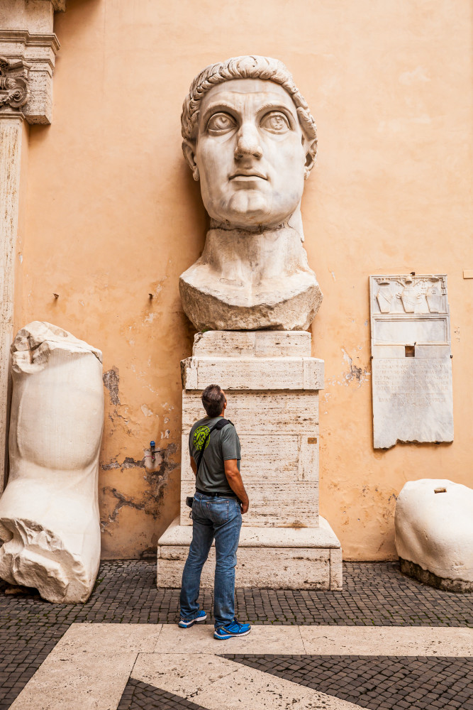 The fragments and head of the colossal statue of Constantine with a man standing below looking up at it. Capitoline Museum, Rome Italy.