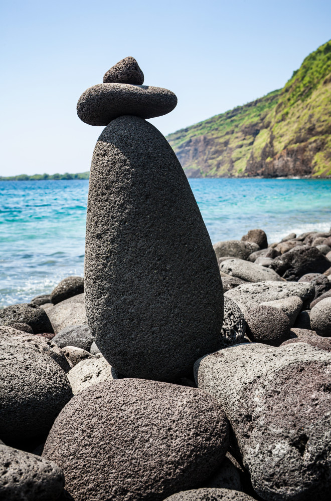 A rock cairn on a rocky beach with Kealakekua Bay in the background and captain Cook Memorial on the opposite side of the bay. Hawaii, South Kona.