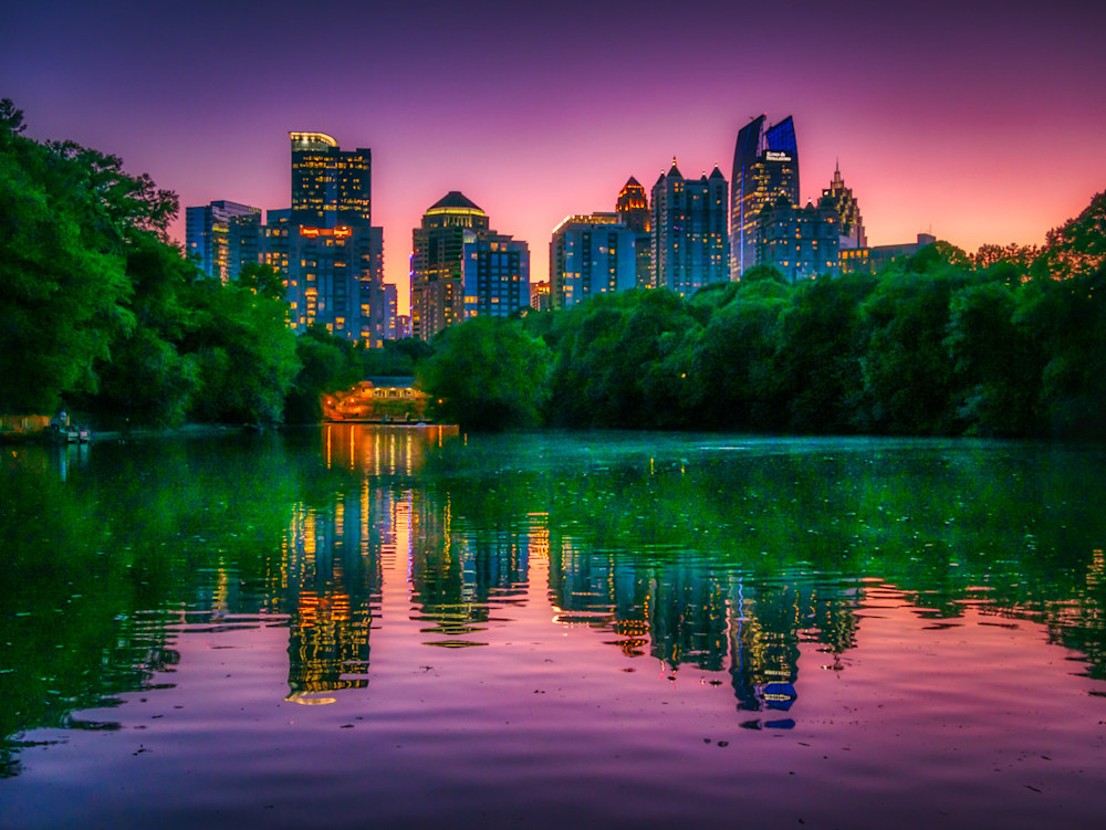 The City of Atlanta as seen from Piedmont Park at night with a saturated purple, pink, and orange sky and bright green leaves on the trees with reflections of the city in Lake Clara Meer