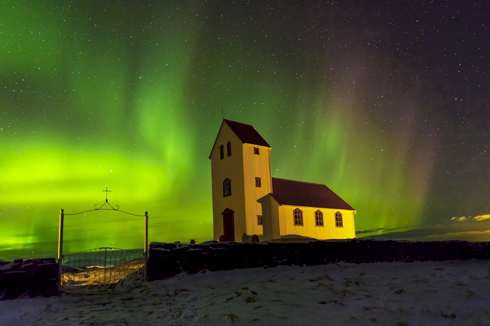 Northern lights glowing green and purple above a white country church beneath a starry sky, evoking mystery and spirit