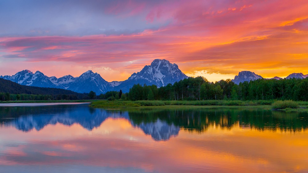 Sunset reflecting in a calm lake beneath the Teton mountains with a vibrant orange, pink, and purple sky