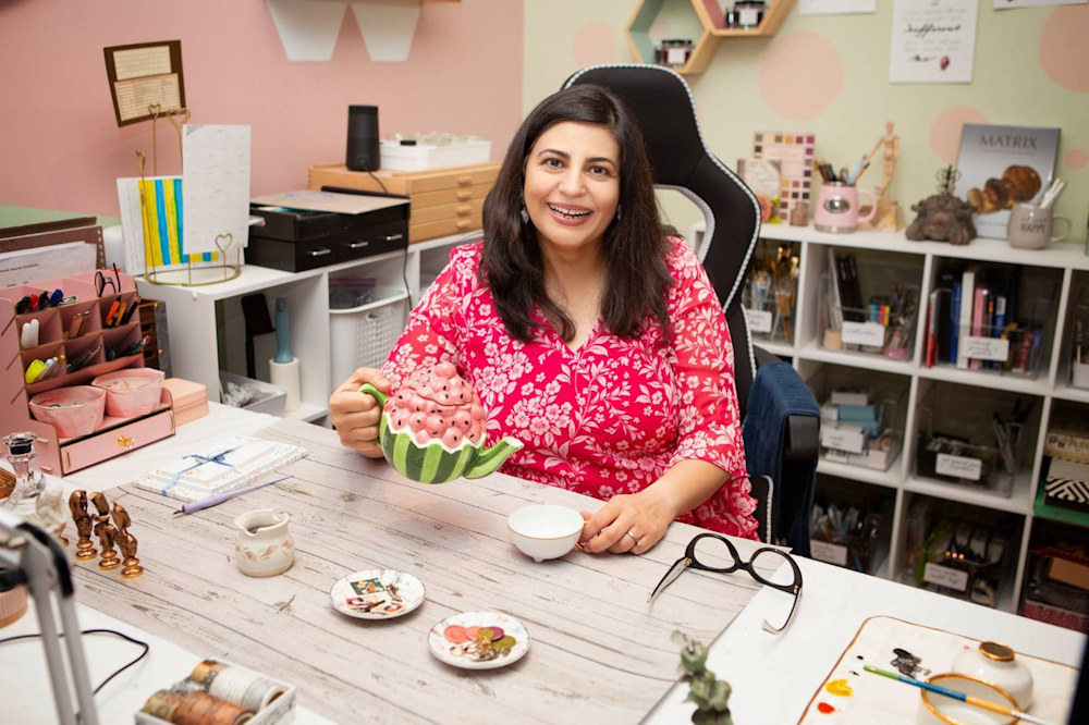 Sonia J. Pal in her art studio, smiling and holding a watermelon-shaped teapot, surrounded by creative supplies