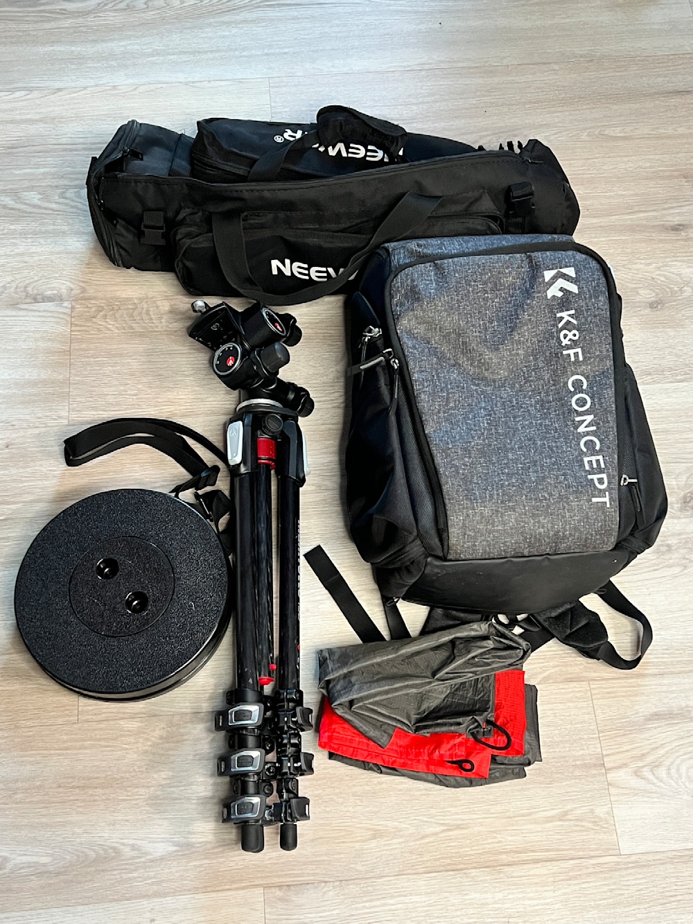 A flat lay of photography gear on a wooden floor, including a black and grey K&F Concept backpack, a black bag, a tripod with a ball head, a black telescopic stool, and a folded waterproof picnic mat.