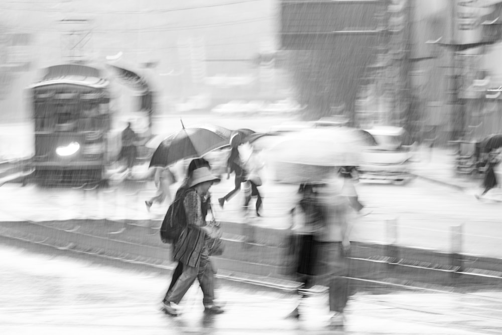Rainy street scene in Kumamoto with umbrellas compressed by motion blur; central figure’s confident stride resembles a runway walk.