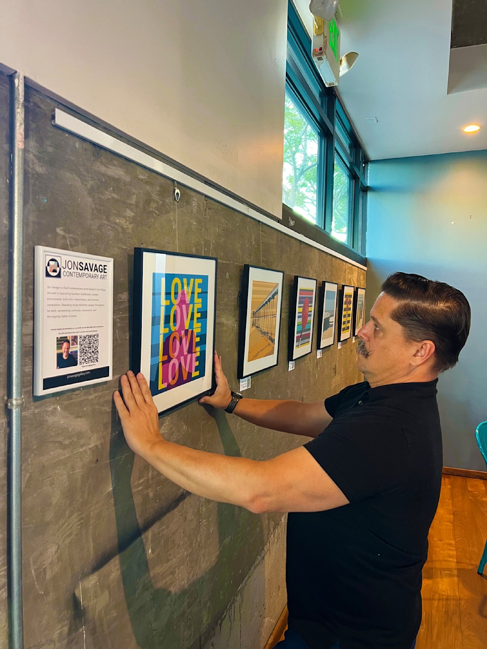 Jon Savage hand-hanging the 'Love 4 More' fine art print on a modern concrete gallery wall at Bird Rock Coffee, 2295 Kettner Blvd.