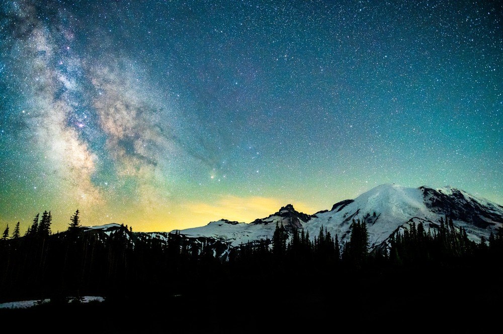 Photo of Mt. Rainier in Washington State at night with a green and blue starry night sky with the milky way on the left.