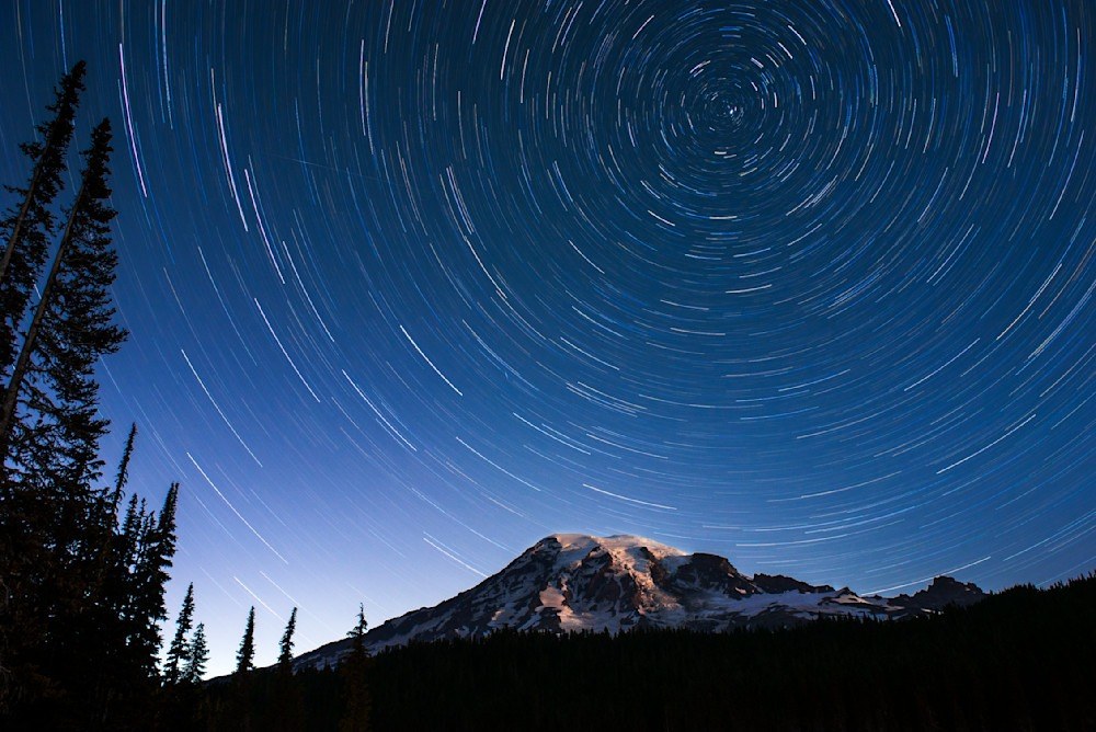 Photo of Mt. Rainier at night with the mountain lit up with ambient white light with a circular trail of stars circling around Polaris above the mountain.